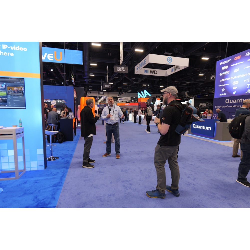 A busy convention floor with several people standing and talking in a large exhibition hall with blue carpeting and multiple branded booths and screens.