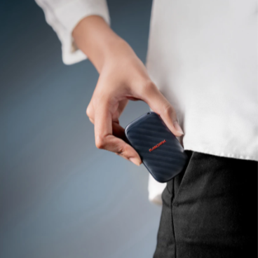 A close-up of a person's hand sliding a small, dark blue-gray rectangular device with a red logo into their white shirt pocket. The person is wearing a white long-sleeve shirt and black trousers, set against a dark blue-gray gradient background.