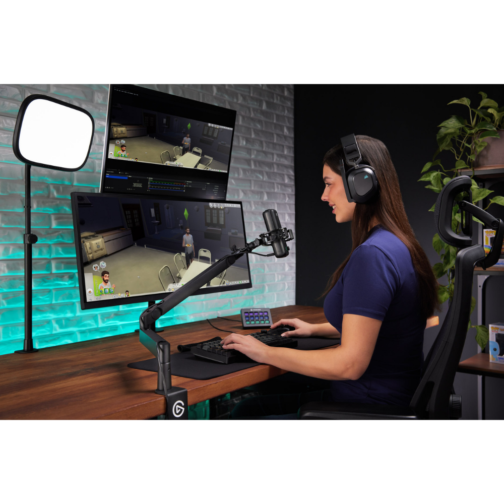 A woman sits at a desk using a dual-monitor gaming setup with an adjustable monitor arm, keyboard, mouse, and headset. A desktop microphone, monitor light panel, and chair are visible in a styled home office environment.
