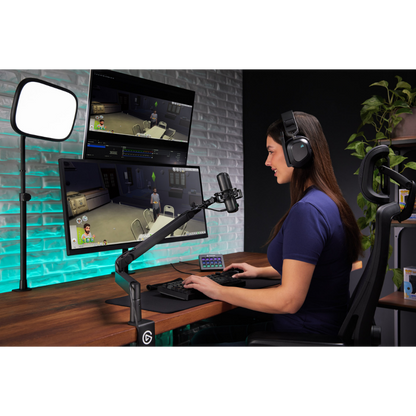 A woman sits at a desk using a dual-monitor gaming setup with an adjustable monitor arm, keyboard, mouse, and headset. A desktop microphone, monitor light panel, and chair are visible in a styled home office environment.