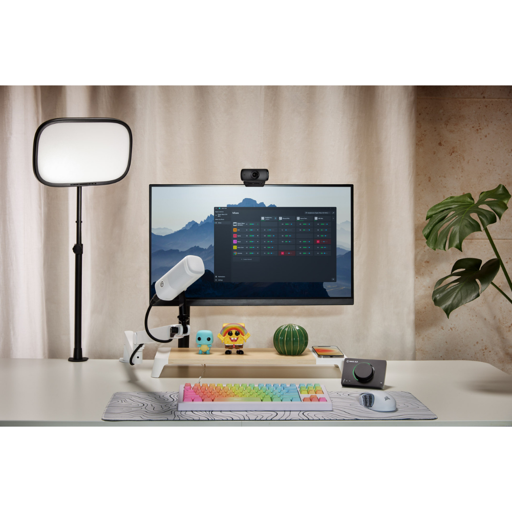 A desktop setup scene showing a computer monitor with a webcam mounted above it, a floor lamp on the left, a desk shelf with small decor items, a keyboard, mouse, and mousepad on a table, and a potted plant on the right. No person is present, but this is not a clean isolated product shot because it is staged in a real interior environment, so subject_type is Lifestyle.