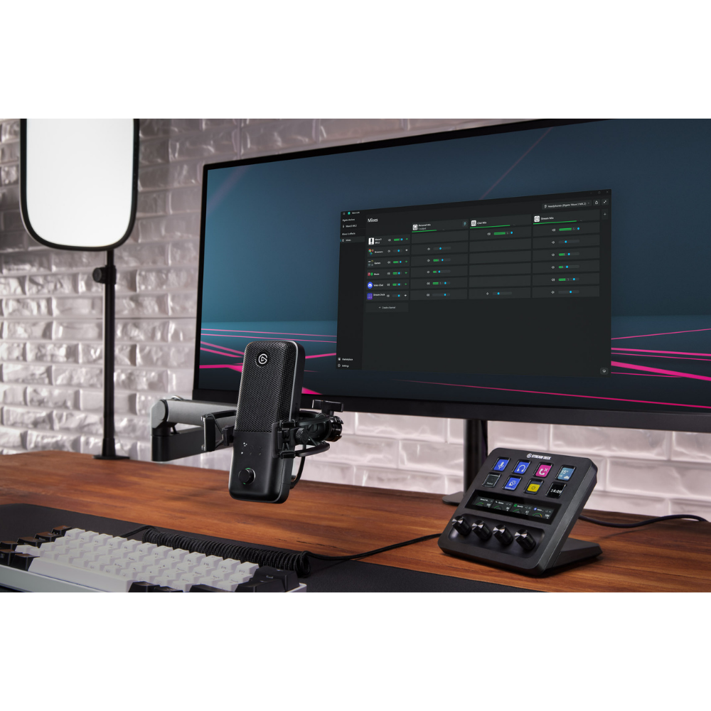 A desktop audio setup featuring a computer monitor, a microphone on an arm, and a control panel with a small display and knobs, arranged on a wooden desk against a light brick wall.