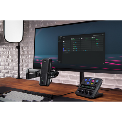 A desktop audio setup featuring a computer monitor, a microphone on an arm, and a control panel with a small display and knobs, arranged on a wooden desk against a light brick wall.