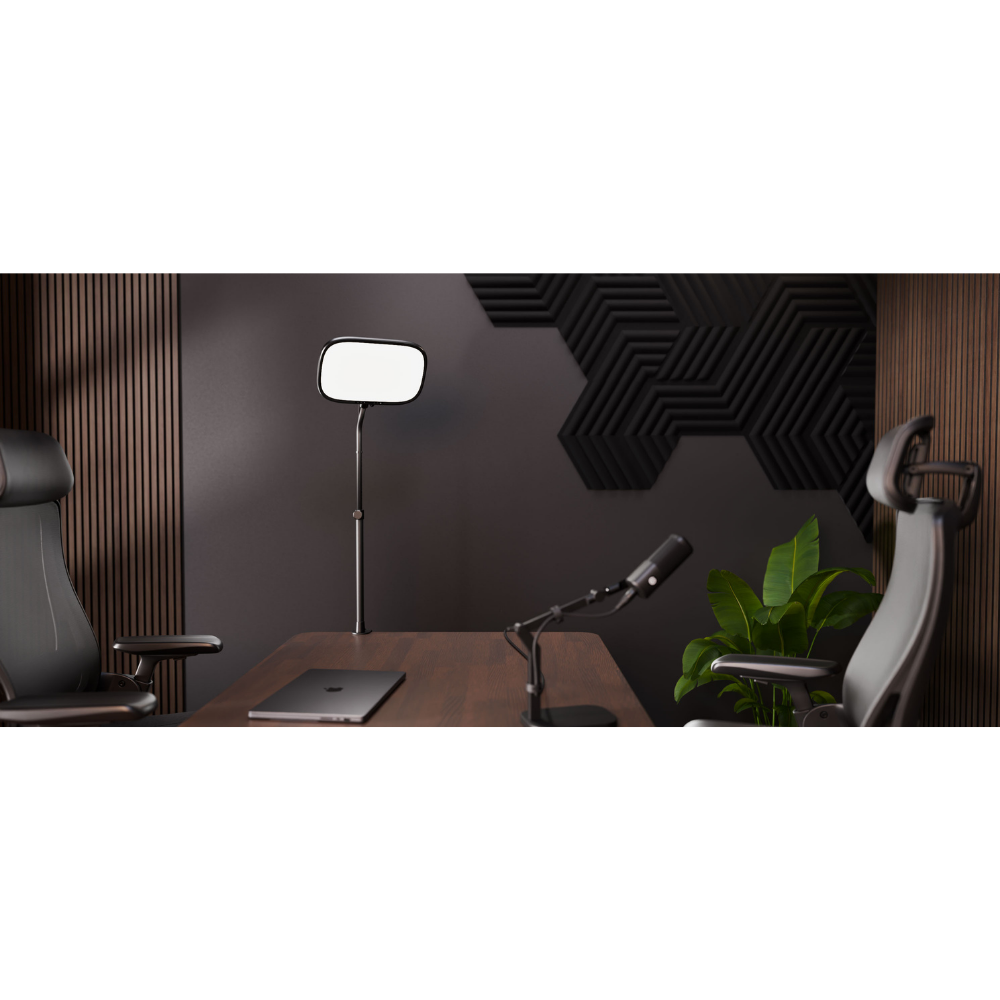 A modern home office setup featuring a dark wood desk, a black standing floor lamp with a rectangular head, a desktop microphone on an adjustable arm, a closed silver laptop, and two ergonomic office chairs against a dark gray wall with geometric acoustic panels.