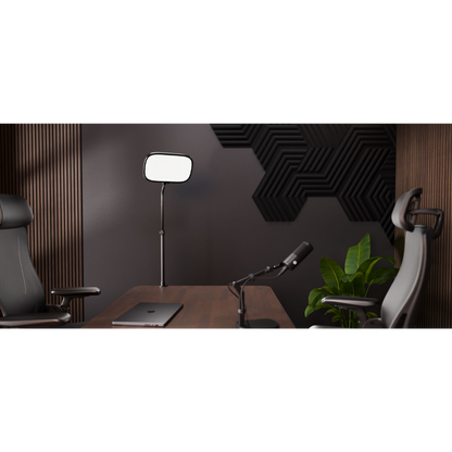 A modern home office setup featuring a dark wood desk, a black standing floor lamp with a rectangular head, a desktop microphone on an adjustable arm, a closed silver laptop, and two ergonomic office chairs against a dark gray wall with geometric acoustic panels.