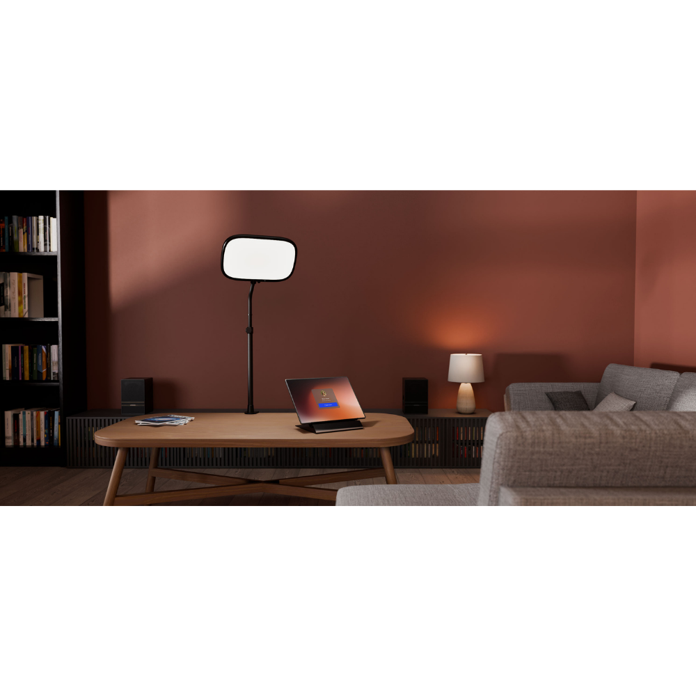 A modern living room scene with a black floor lamp featuring a rectangular white diffuser panel, styled on a wooden coffee table next to an open laptop. The background is a warm terracotta wall with a gray sofa and bookshelves.