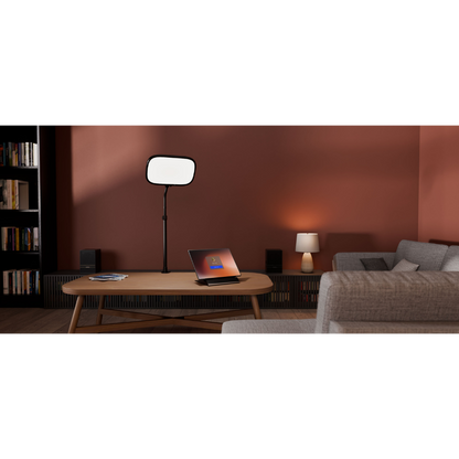 A modern living room scene with a black floor lamp featuring a rectangular white diffuser panel, styled on a wooden coffee table next to an open laptop. The background is a warm terracotta wall with a gray sofa and bookshelves.