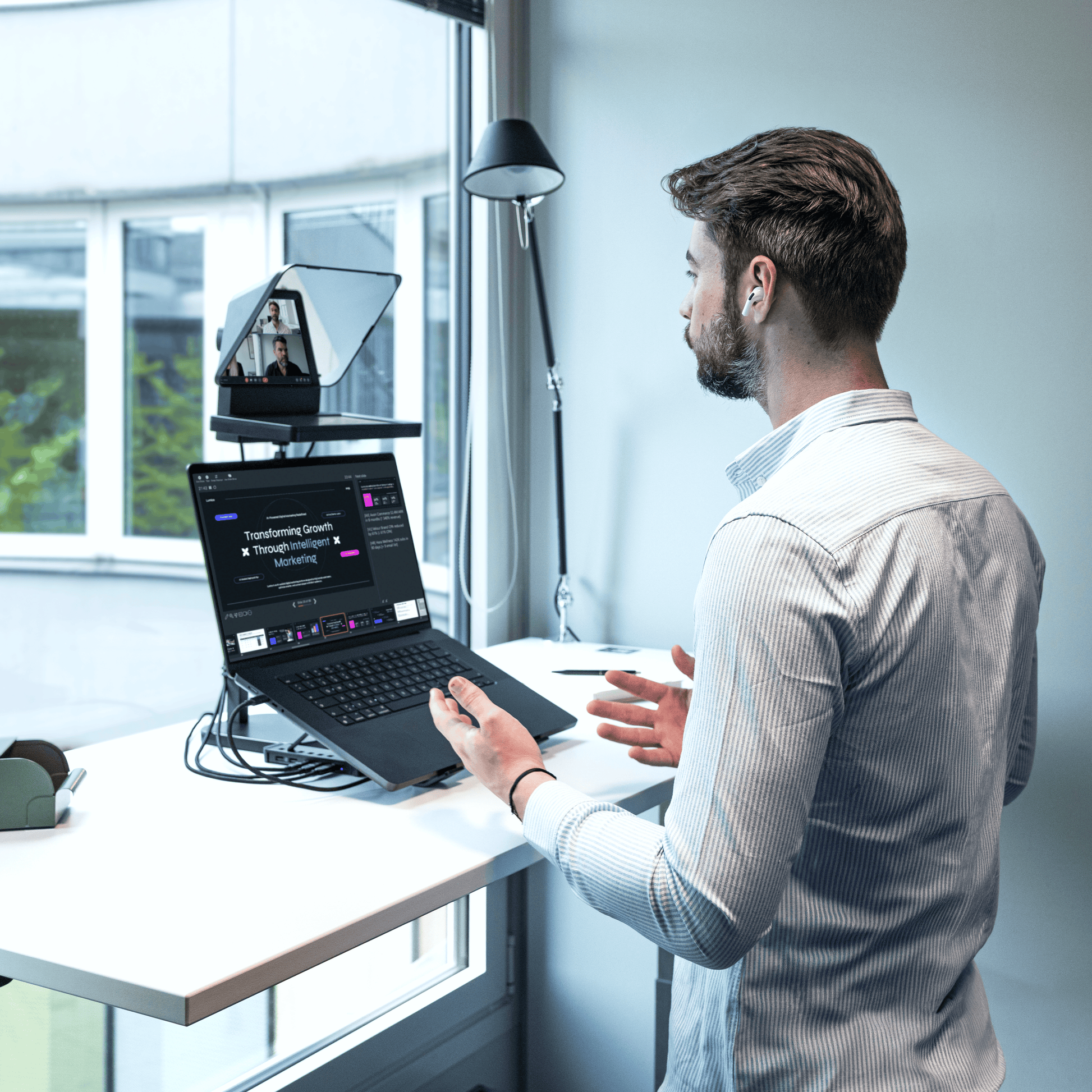 A man in a light gray striped shirt stands at a white desk in a bright office, using a laptop with a mounted webcam and microphone setup. The scene is lit by natural light from a large window, with a black floor lamp in the background.