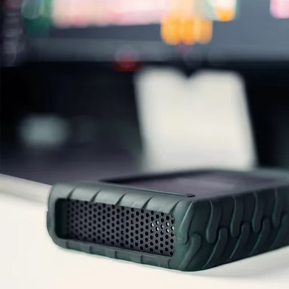 A rugged black portable speaker with a textured rubberized bumper and a perforated grille, shown in a close-up shot on a white surface with a blurred background.