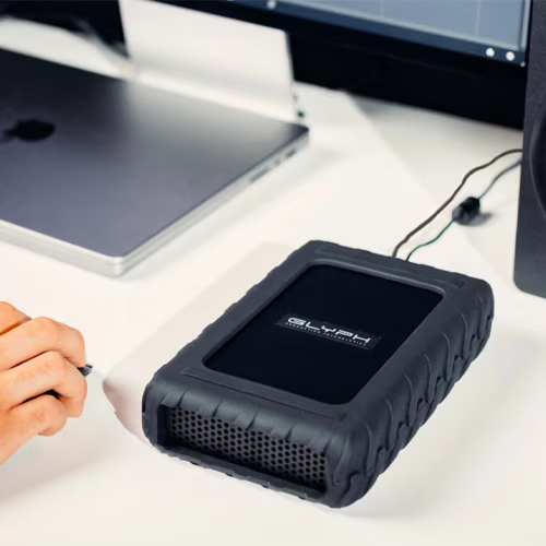 A rugged black external hard drive by GLYPH on a white desk, with a hand holding a pen nearby and a laptop in the background.