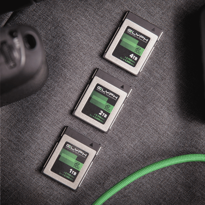 Three Glyph CFexpress memory cards of different capacities on a textured surface with a green cable.