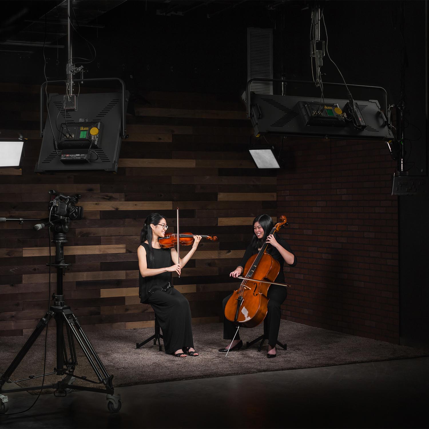 Two musicians playing violin and cello in a dark studio with lighting equipment.