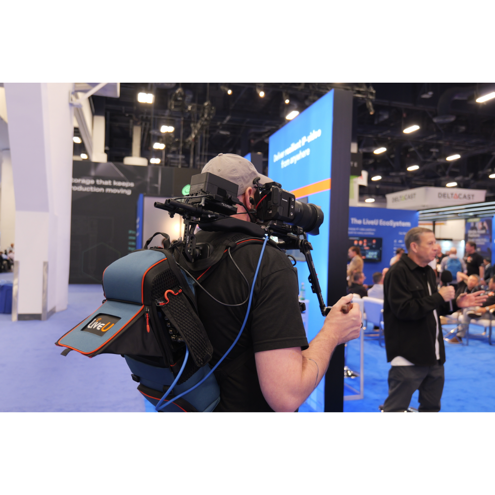 A person at a conference wearing a backpack-mounted camera rig with a large shoulder mount, shoulder rest, and side storage pouch. The setup includes visible cables and a handheld remote, photographed in a real event hall with booths and other people in the background.