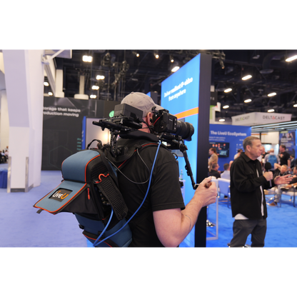 A person at a conference wearing a backpack-mounted camera rig with a large shoulder mount, shoulder rest, and side storage pouch. The setup includes visible cables and a handheld remote, photographed in a real event hall with booths and other people in the background.