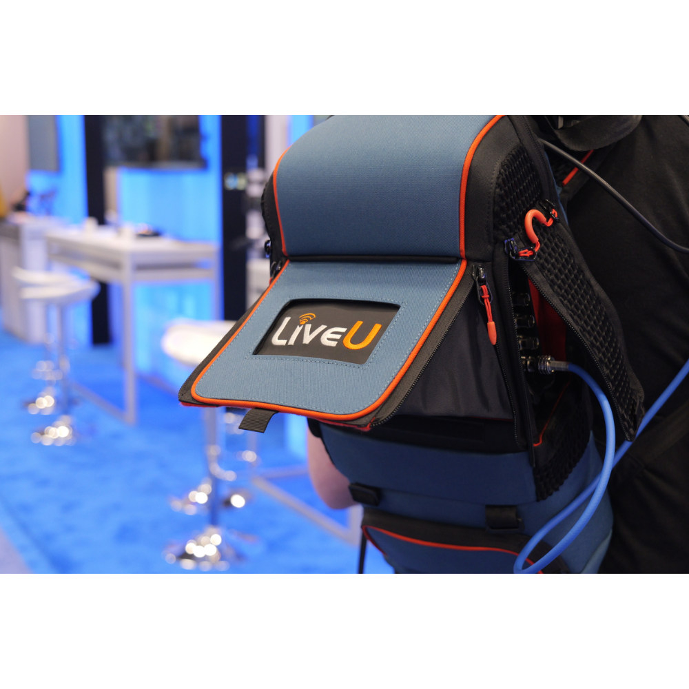 A close-up of a blue and black LiveU backpack with orange trim, worn by a person in a convention or exhibition setting with a blue carpeted floor and blurred booths in the background.