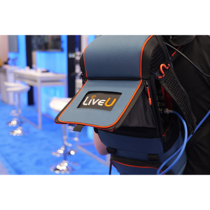 A close-up of a blue and black LiveU backpack with orange trim, worn by a person in a convention or exhibition setting with a blue carpeted floor and blurred booths in the background.