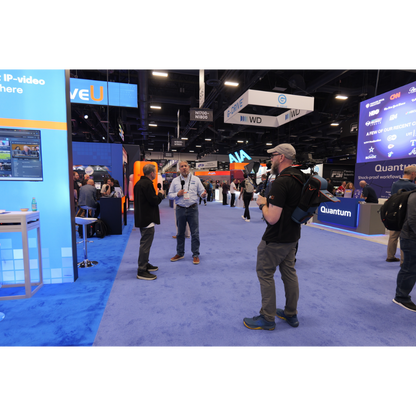 A busy convention floor with several people standing and talking in a large exhibition hall with blue carpeting and multiple branded booths and screens.