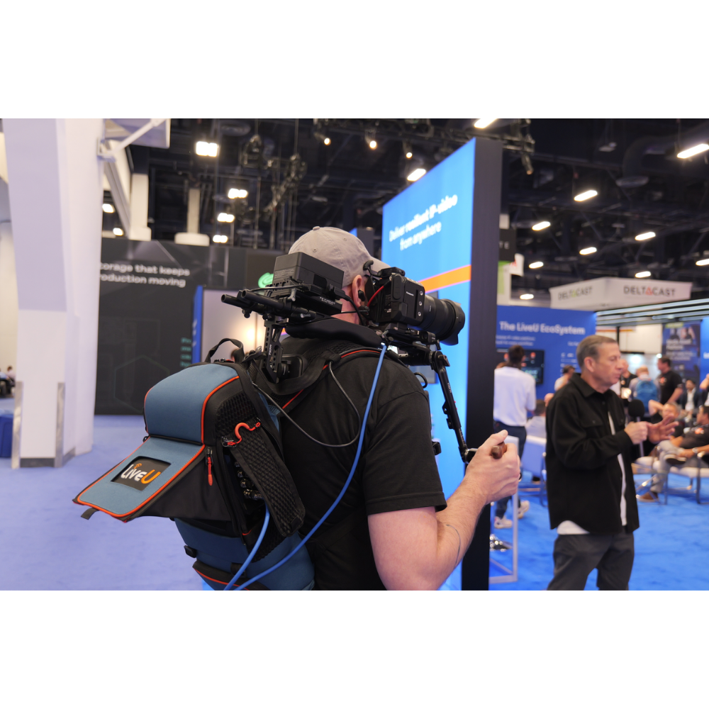 A man at a trade show or conference wearing a backpack-mounted video camera rig on his back, seen from behind in a large indoor exhibition hall with booths and blue carpeting.