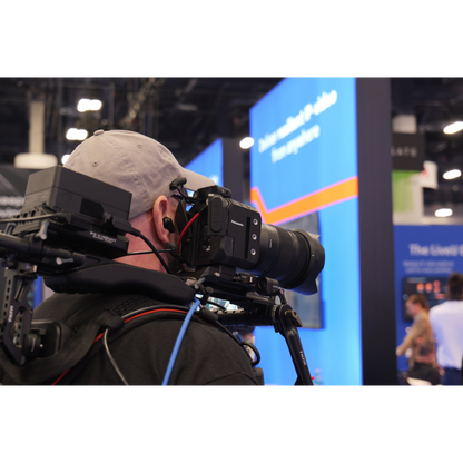 A person wearing a light gray cap and dark clothing is shown from the side, holding a large black video camera with an attached lens in a busy indoor event or exhibition hall. Blue signage and other people are visible in the background.