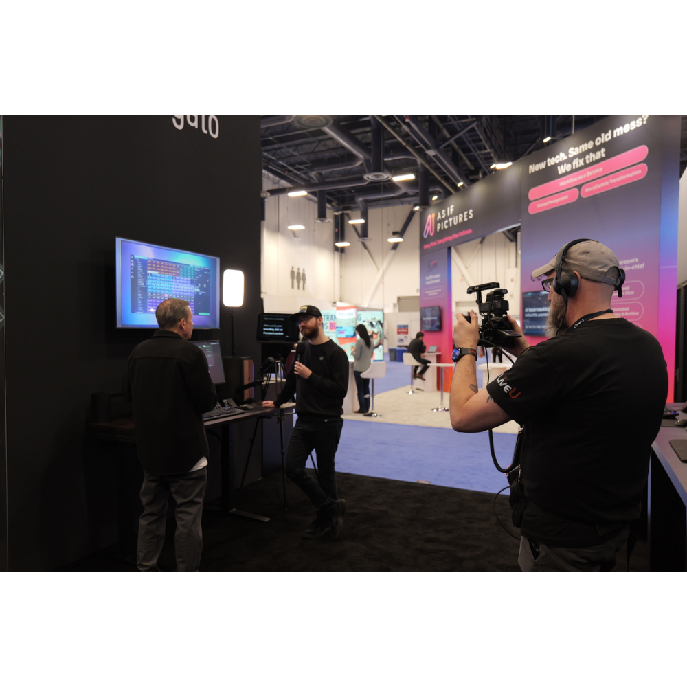 A conference or exhibition hall scene with several people around a display booth. A cameraman in the foreground holds a video camera, while others stand near a screen and booth setup.