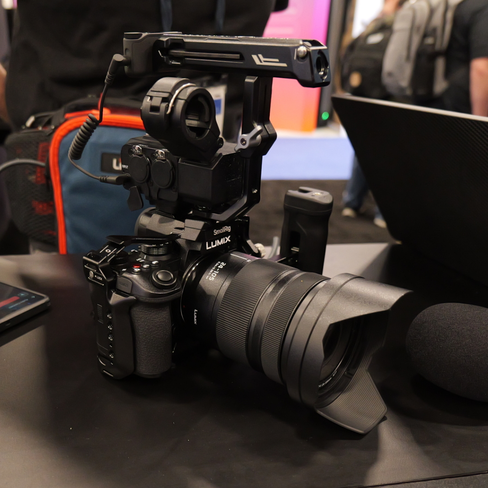 A black Panasonic Lumix camera with a large lens, mounted on a rig with a scope and handle, sitting on a dark table with a blurred background.