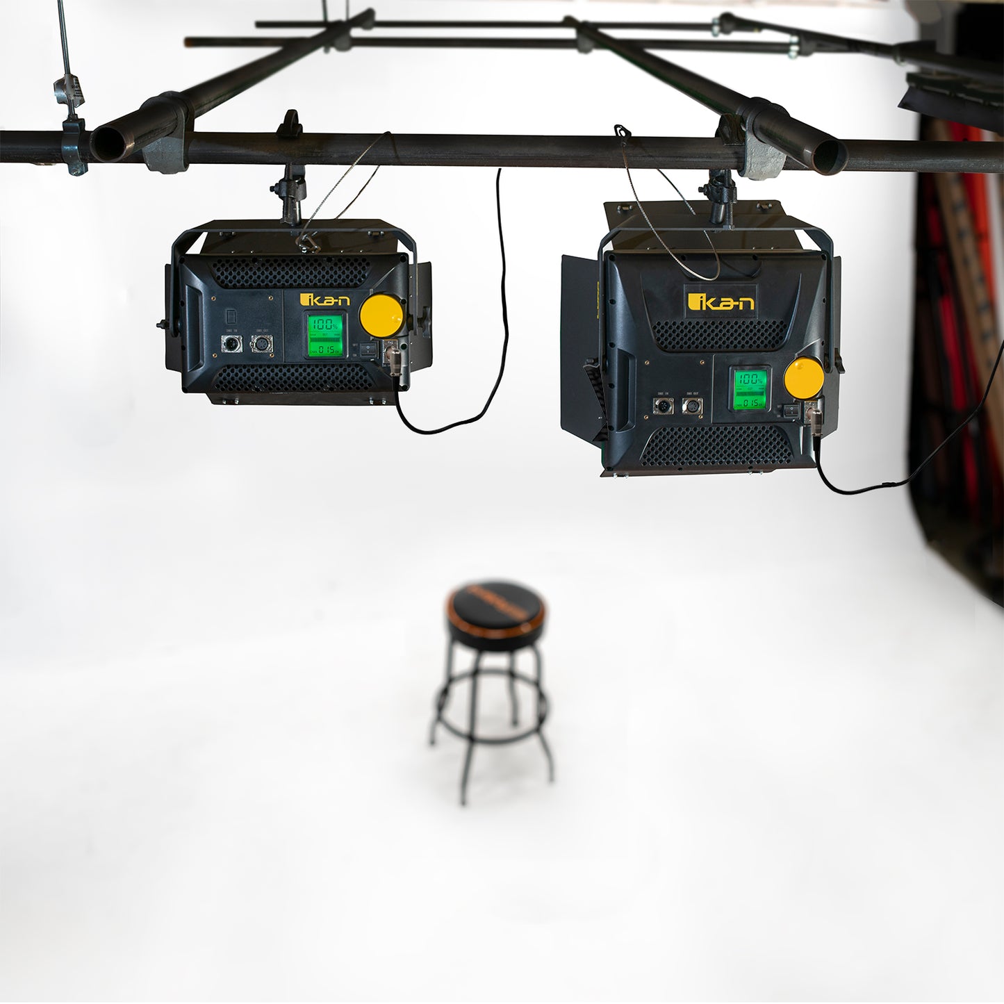 Two black electronic devices with yellow buttons hanging from a bar above a stool on a white background.