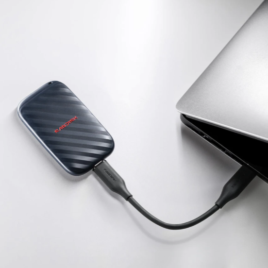 A black external hard drive with a red 'SANDISK' logo, connected via a black USB cable to a silver laptop on a clean white surface.