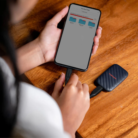 A person holds a smartphone connected to a small black portable SSD on a wooden table. The SSD has a textured dark gray surface with a small red logo that appears to read STORMER.